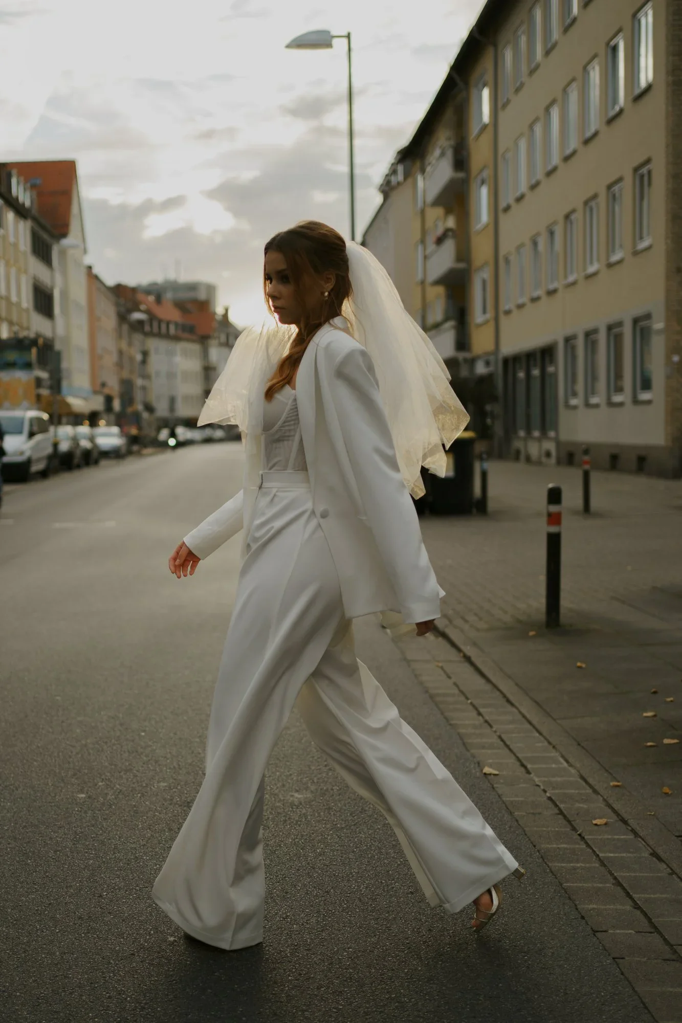 Eine Frau in weißer Brauthose und Corsage überquert eine Straße. Sie trägt einen Hochzeitsschleier auf dem Kopf.