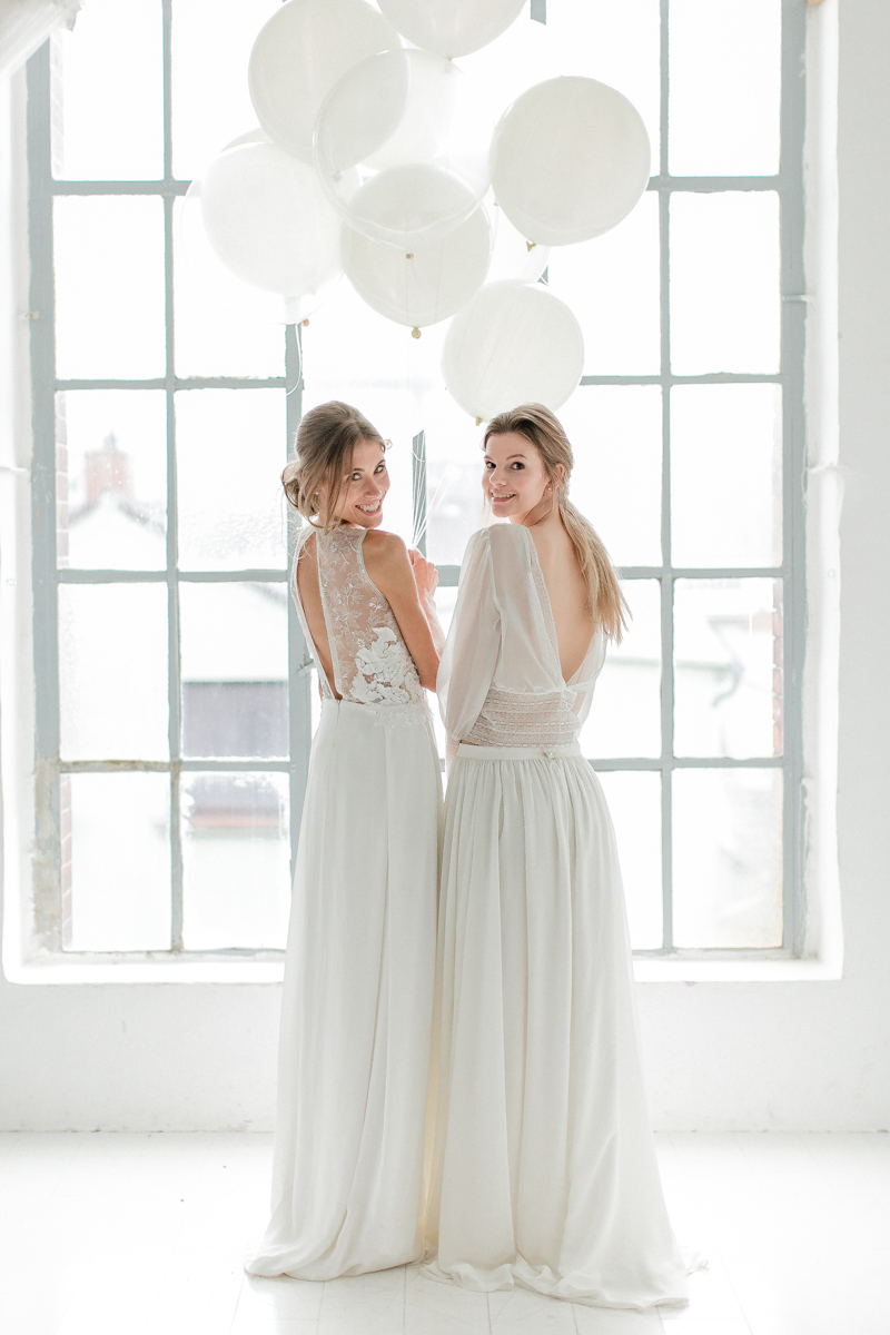 Zwei Bräute in Hochzeitskleidern stehen in einem hellen Raum und halten weiße Luftballons in den Händen