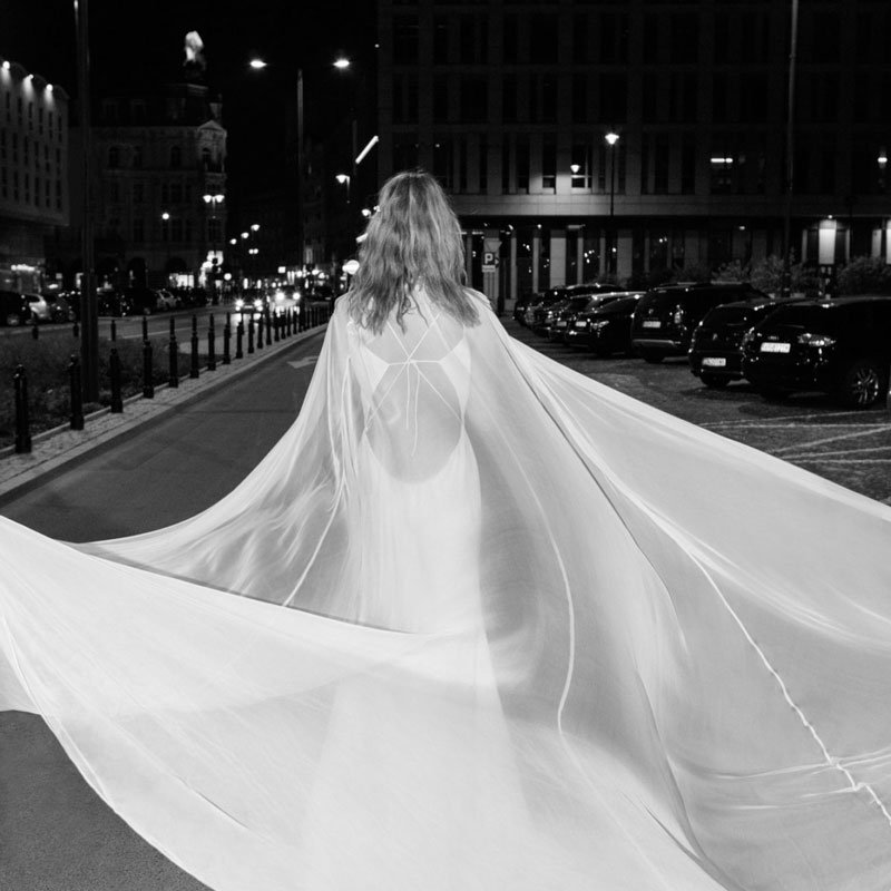 Eine Braut im weißen Brautkleid und einem wehenden Schleier steht mit dem Rücken zur Kamera nachts auf einer Straße.