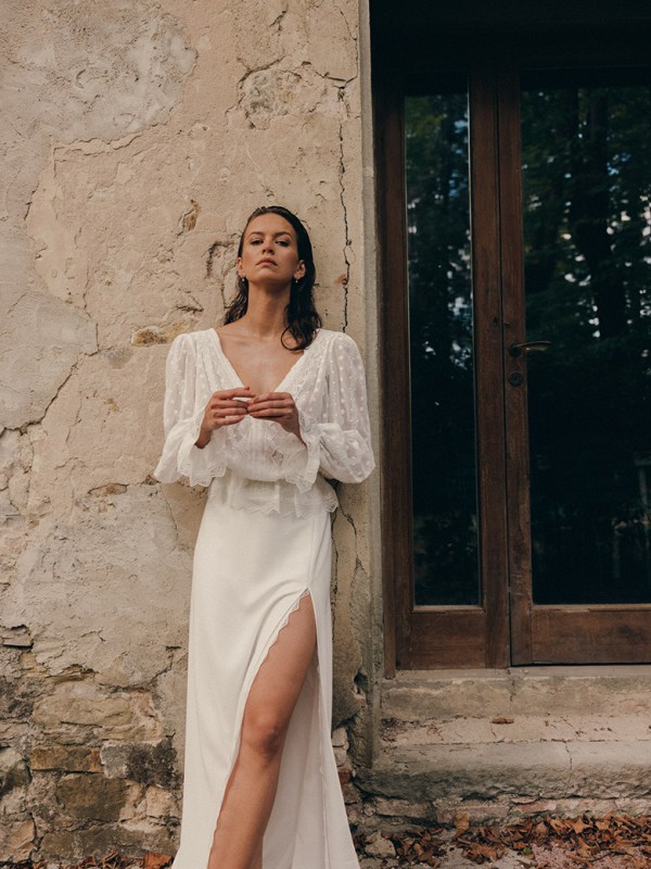 Brautkleid mit langem Ärmel aus zartem Pünktchentüll und tiefem V Ausschnitt mit hohem Beinschlitz vor einer verwitterten Hauswand