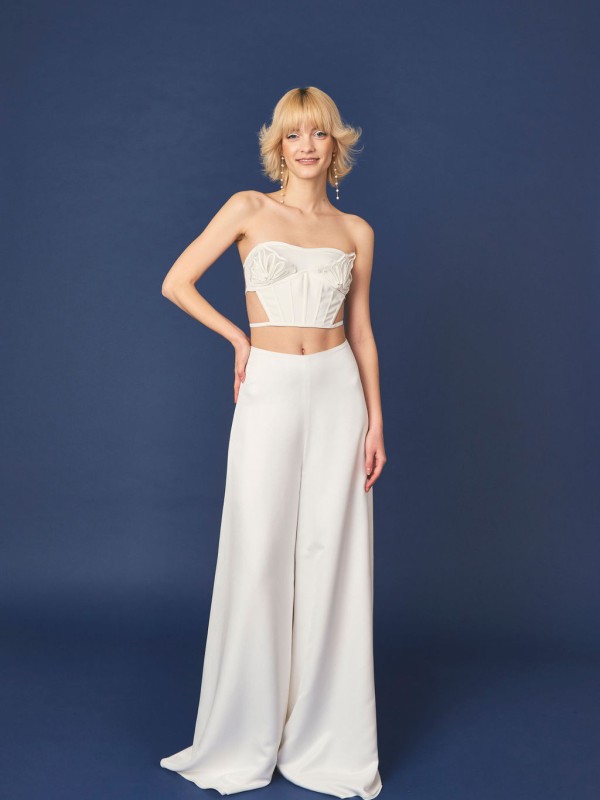 Moderner Jumpsuit für die Hochzeit mit trägerlosem Bustier Oberteil und weit fallender weißer Hose vor dunkelblauem Hintergrund