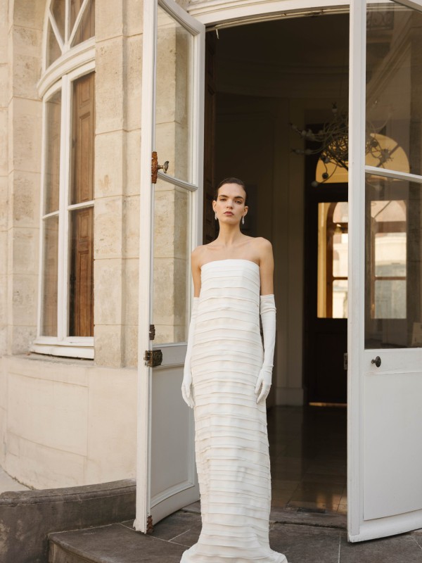 Plissiertes, schulterfreies Brautkleid in gerader Silhouette mit langen weißen Handschuhen, getragen vor einem klassischen Gebäude