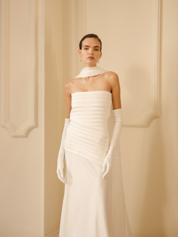 Elegantes, klassisches Brautkleid mit plissiertem Oberteil und geradem Ausschnitt, kombiniert mit langen weißen Handschuhen und Chiffon-Schal vor heller Wandverzierung