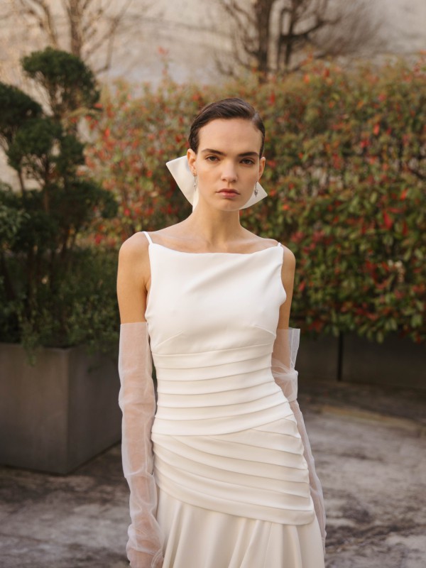 Klassisches Brautkleid mit Spaghettiträgern, plissiertem Taillenbereich und transparenten Handschuhen, getragen im eleganten Außenbereich mit grün-roter Bepflanzung