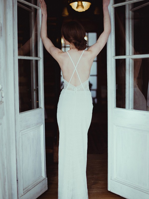 Boho-inspiriertes rückenfreies Brautkleid aus fein gemustertem Spitzenstoff mit schmalen überkreuzten Spaghettiträgern, Braut steht mit erhobenen Armen in weißem Türrahmen vor dunkler Kulisse