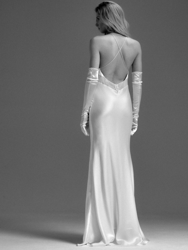 Elegantes rückenfreies Brautkleid aus Satin mit schmalen Spaghettiträgern und langen Handschuhen, Braut mit langen Haaren und klassischem Look in Schwarzweiß vor schlichtem Hintergrund