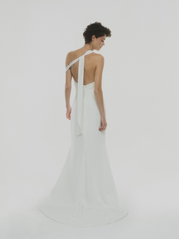 Modernes rückenfreies Brautkleid mit asymmetrischem Träger und langer Stoffschleife, schlicht und elegant, Braut mit kurzer dunkler Lockenfrisur vor hellem Hintergrund