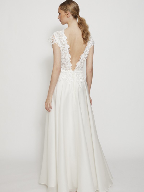 Elegantes rückenfreies Brautkleid mit floraler Spitze, kurzen Ärmeln und tiefem V-Ausschnitt am Rücken, weiter Rock aus fließendem Stoff, Braut mit blondem Zopf vor hellem Hintergrund