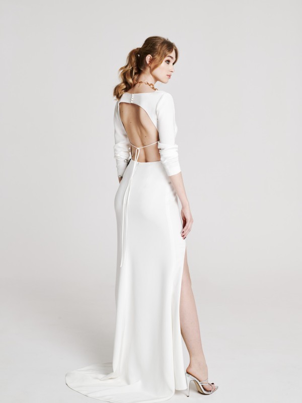 Modernes rückenfreies Brautkleid mit raffiniertem Cut-Out und feinem Bindeband im Rücken, langem Beinschlitz, elegante Braut mit blondem Zopf und hohen Schuhen vor hellem Studiohintergrund