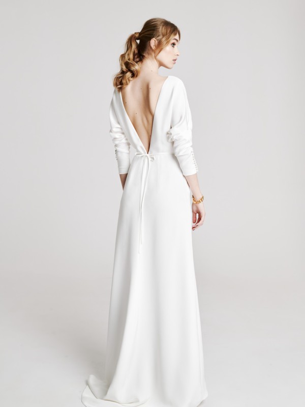 Modernes rückenfreies Brautkleid mit tiefem V-Ausschnitt am Rücken, langen Ärmeln und feinem Bindegürtel, eleganter Look, Braut mit blondem Zopf vor hellem Hintergrund