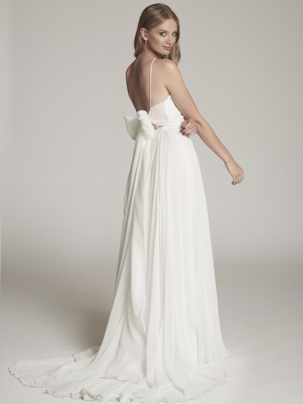 Romantisches rückenfreies Brautkleid mit schmalen Trägern und großer Schleife im Rücken, fließender Stoff, Braut mit langen blonden Haaren vor hellem Hintergrund