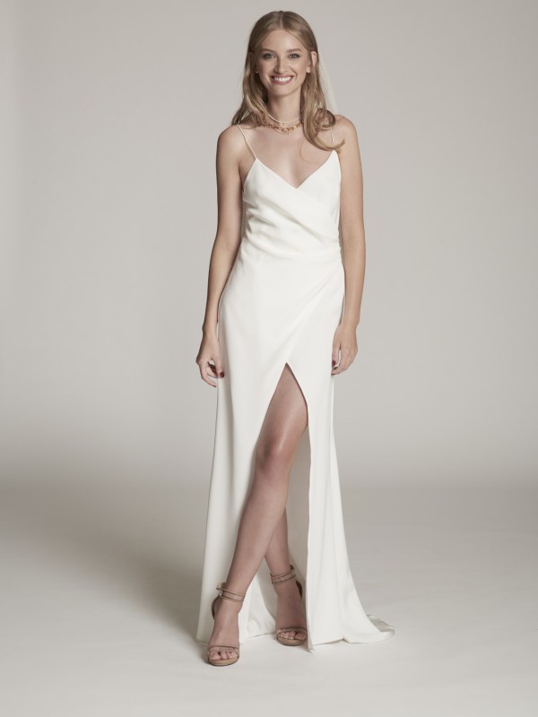 Clean-Brautkleid mit schmalen Spaghettiträgern, Wickeloptik und hohem Beinschlitz, moderne schlichte Silhouette, Braut mit offenem blondem Haar vor hellem Hintergrund