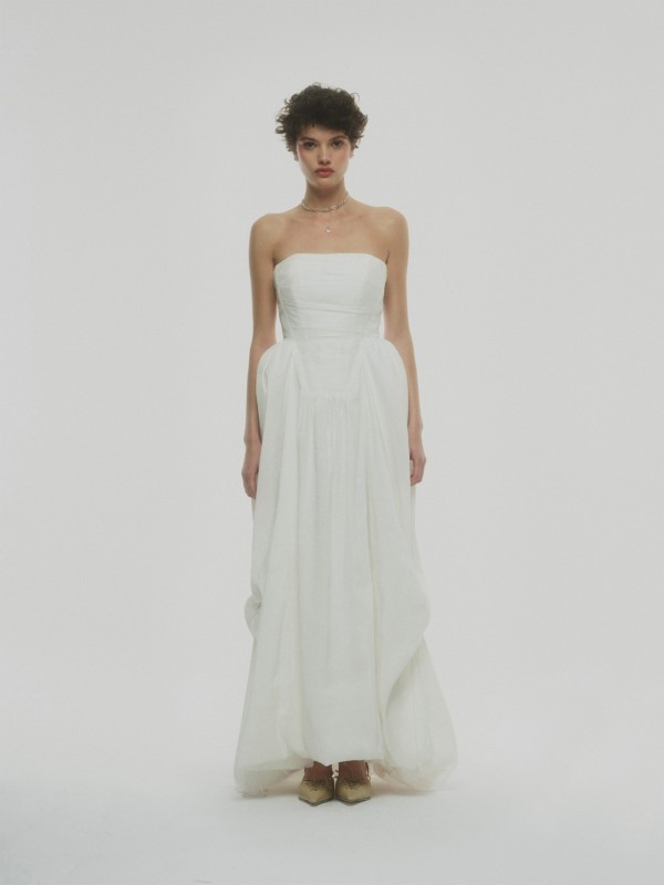 Clean-Brautkleid trägerlos mit voluminösem Rock, schlichtes Design und zarte Taillierung, Braut mit kurzem Haar und Goldschuhen vor weißem Hintergrund