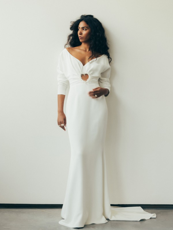 Schlichtes Brautkleid mit langem Ärmel und herzförmigem Cutout in der Taille vor einer weißen Wand