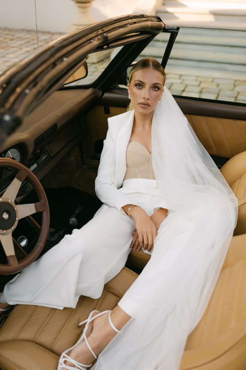 Eine Braut in einem in einem Hosenanzug und lenger Schleppe steht vor einer Villa