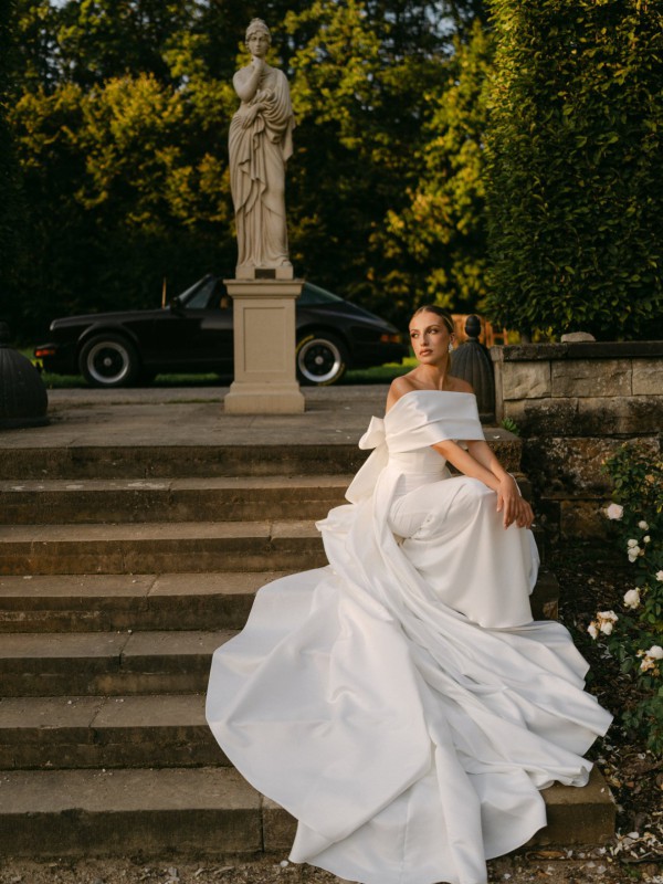 Klassisches Brautkleid mit Schleppe und schulterfreiem Schnitt, fotografiert im Garten mit Statue und Oldtimer im Hintergrund