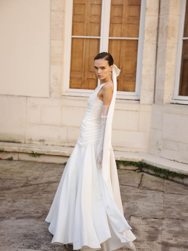 Klassisches Brautkleid mit breiten Trägern, angelegten Falten am Oberteil und langer Schleife im Haar vor einem historischen Gebäude