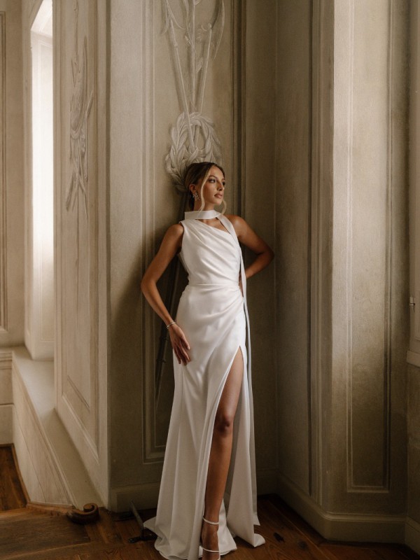 Ein Model steht in einer Villakulisse in einem langen weißen One Shoulder Brautkleid mit Beinschlitz.