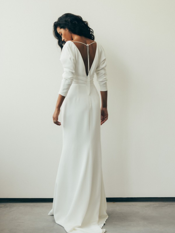 Modernes rückenfreies Brautkleid mit langen Ärmeln, tiefem V-Ausschnitt am Rücken und klarer, schlichter Silhouette, Braut steht vor heller Wand auf grauem Boden