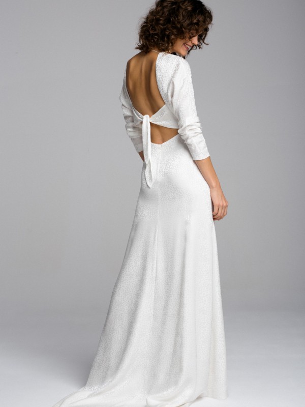 Weißes rückenfreies Brautkleid mit langen Ärmeln und Bindeband am offenen Rückenausschnitt, Braut blickt lächelnd über die Schulter in hellem Studio