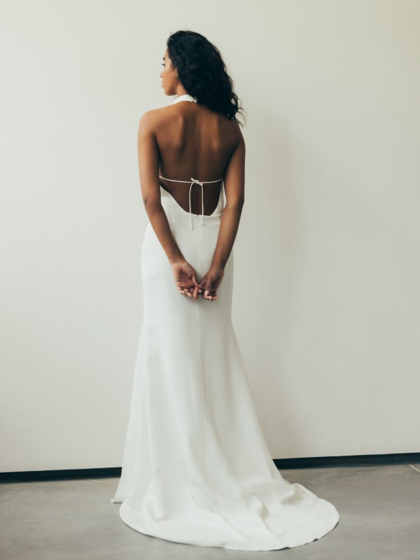 Minimalistisches rückenfreies Brautkleid mit Neckholder und Bindebändern am Rücken, figurbetontes Design, Braut steht mit verschränkten Händen vor heller Wand