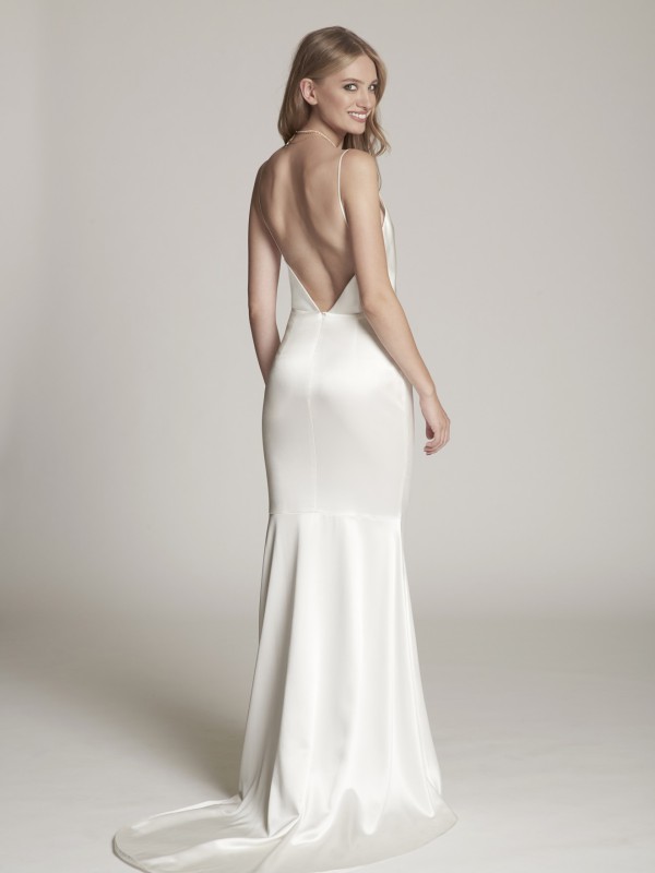 Elegantes rückenfreies Brautkleid aus glänzendem Satin mit tiefem V-Ausschnitt und schmalen Trägern, Braut lächelt über die Schulter vor neutralem Hintergrund