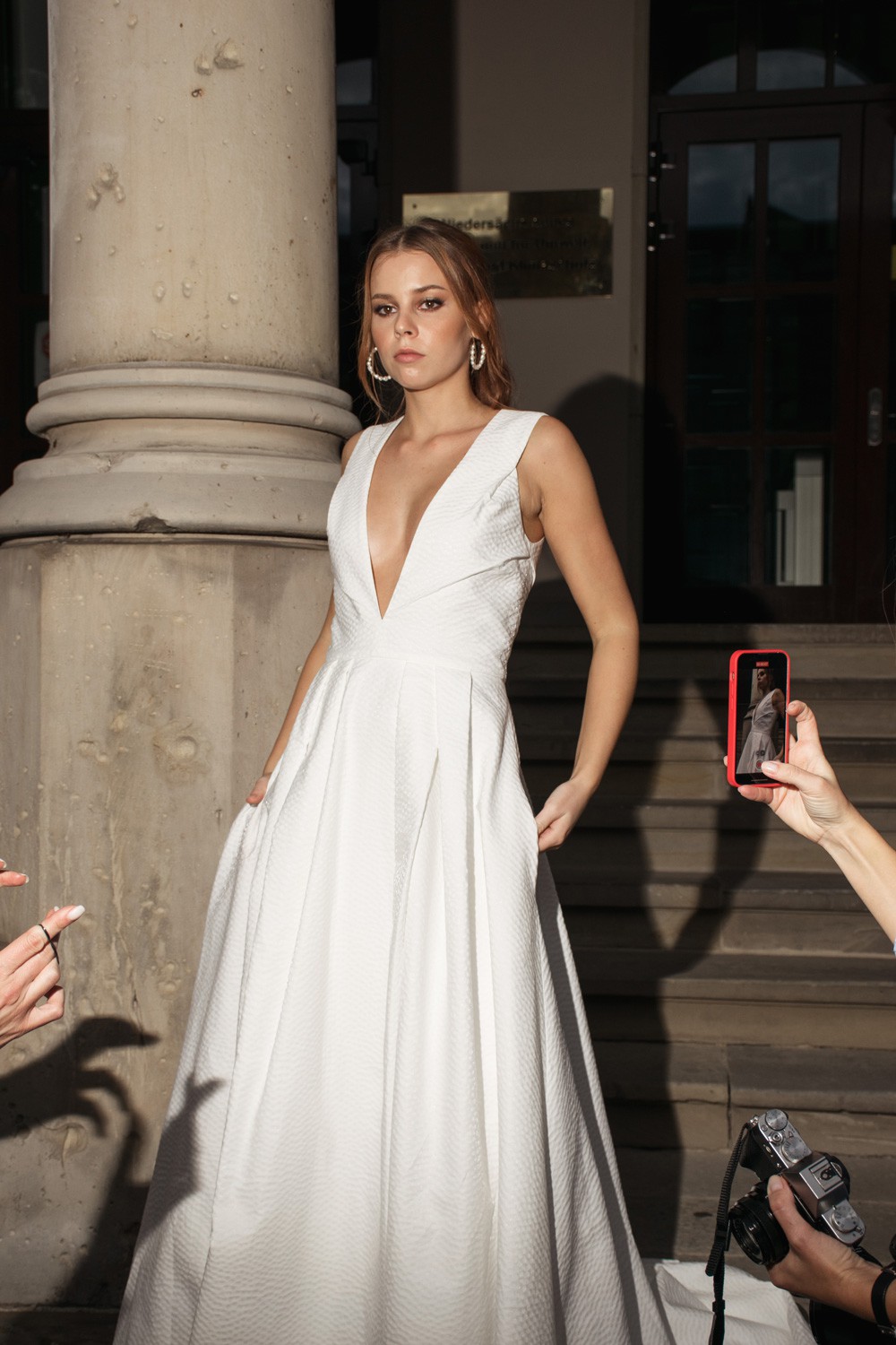 Braut in weißem Kleid mit tiefem Ausschnitt vor einer historischen Säule in der Innenstadt von Hannover, abends mit Fotografenszene.