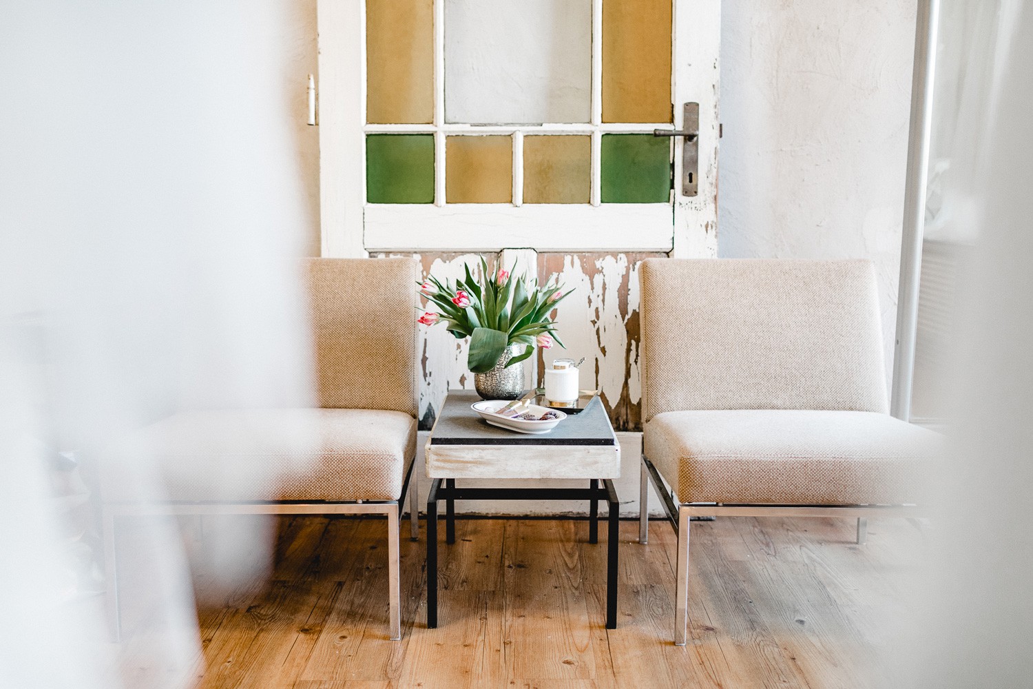 Zwei helle Sessel und ein kleiner Tisch mit frischen Tulpen im Inneren des COSMOPOLKA Bridal Store Hannover, vor einer historischen Tür mit bunten Glaseinsätzen.