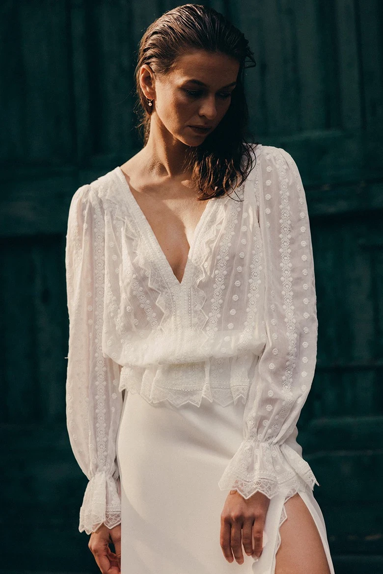 Model in weißem Zweiteiler mit transparenter Bluse und Rock vor dunklem Hintergrund.