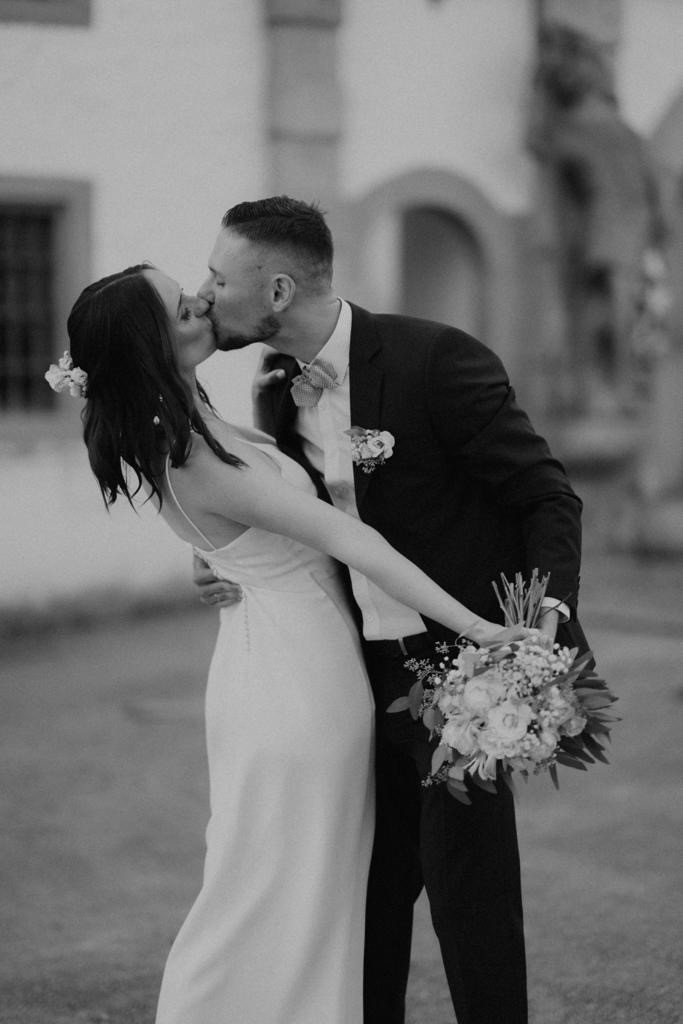Stilvolle Hochzeitsfotografie – Real Bride Anna küsst ihren Bräutigam in einem schlichten, eleganten Brautkleid. Schwarz-weiß-Aufnahme, aufgenommen von Kate Flora Photography.