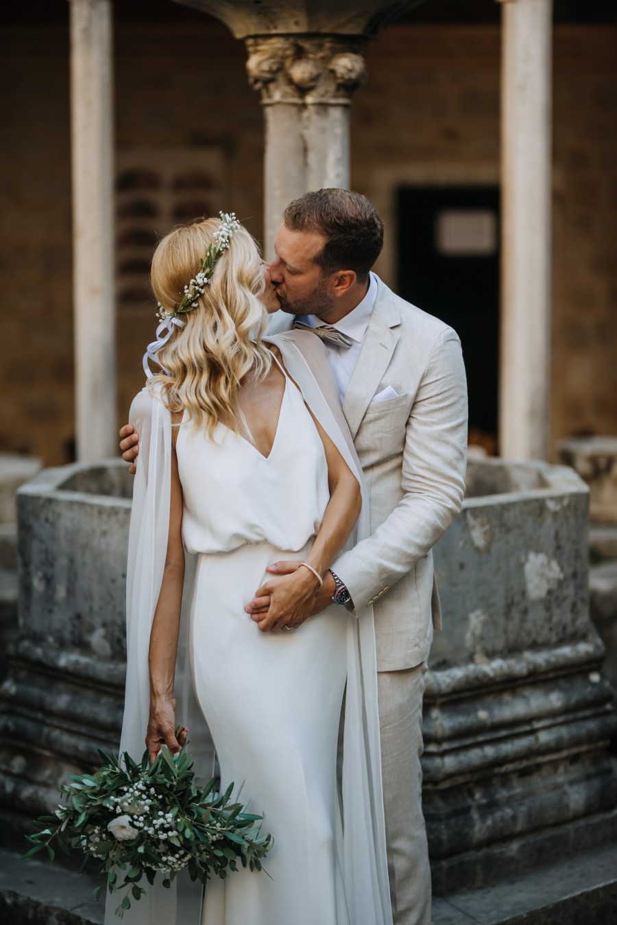 Ein Brautpaar steht vor einem historischen Brunnen und küsst sich. Die Braut hät einen Blumenstrauß in der Hand, der Bräutigam trägt einen weißen Anzug.