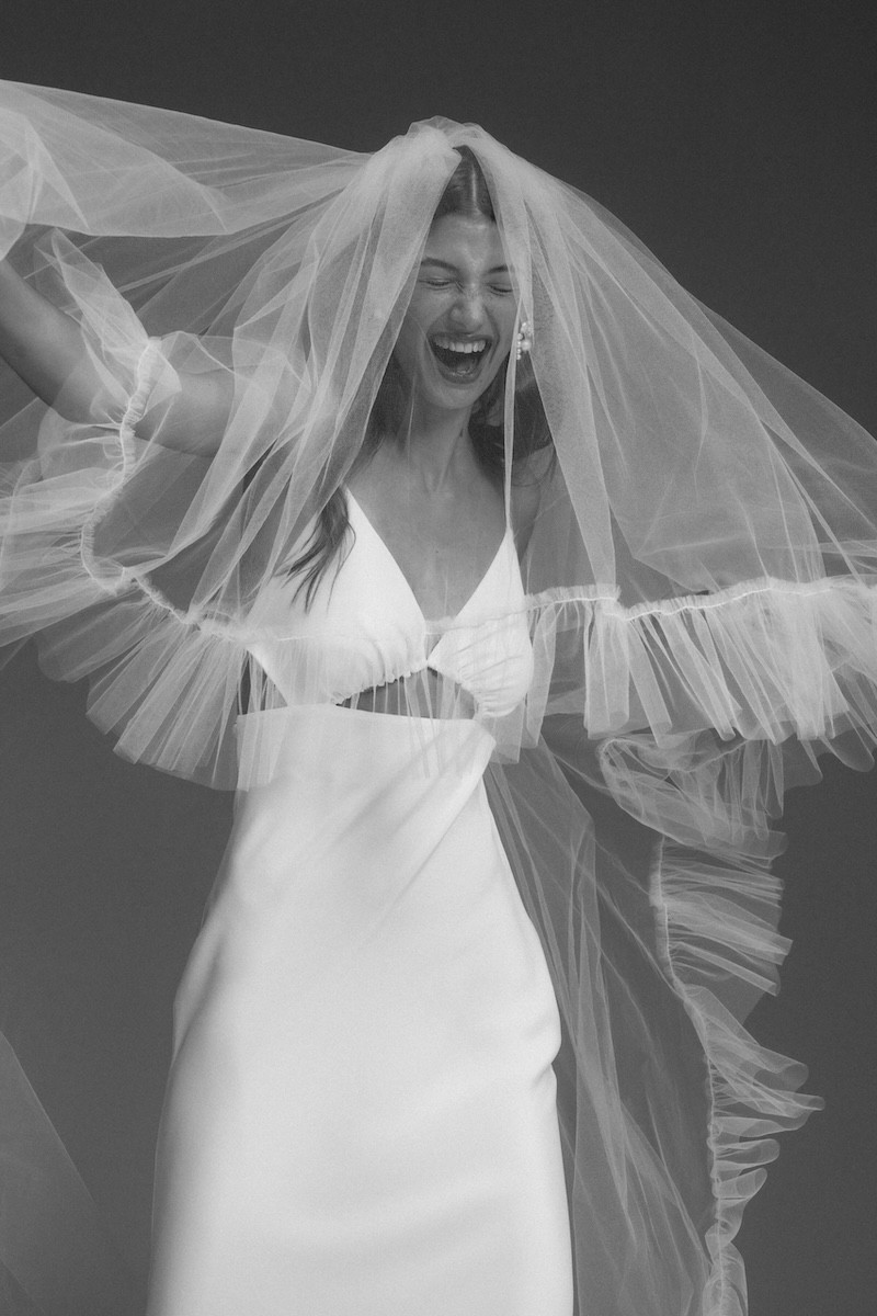 Verspieltes Designer-Brautkleid mit modernen Details, kombiniert mit einem voluminösen, transparenten Schleier. Die Braut strahlt vor Freude, fotografiert in einer Schwarz-Weiß-Ästhetik, die die Leichtigkeit und Eleganz des Looks unterstreicht.