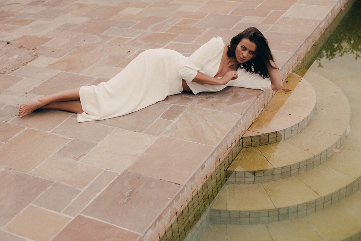 Eine barfüßige Frau im langen Hochzeitskleid liegt auf einem steinboden am Ufer eines Teiches.