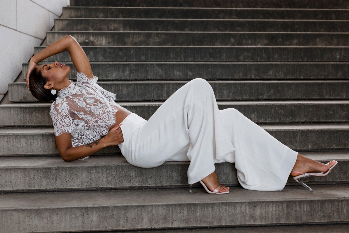 Eine Braut trägt ein durchsitiges Oberteil und weiße Hose und posiert liegend auf einer Treppe.
