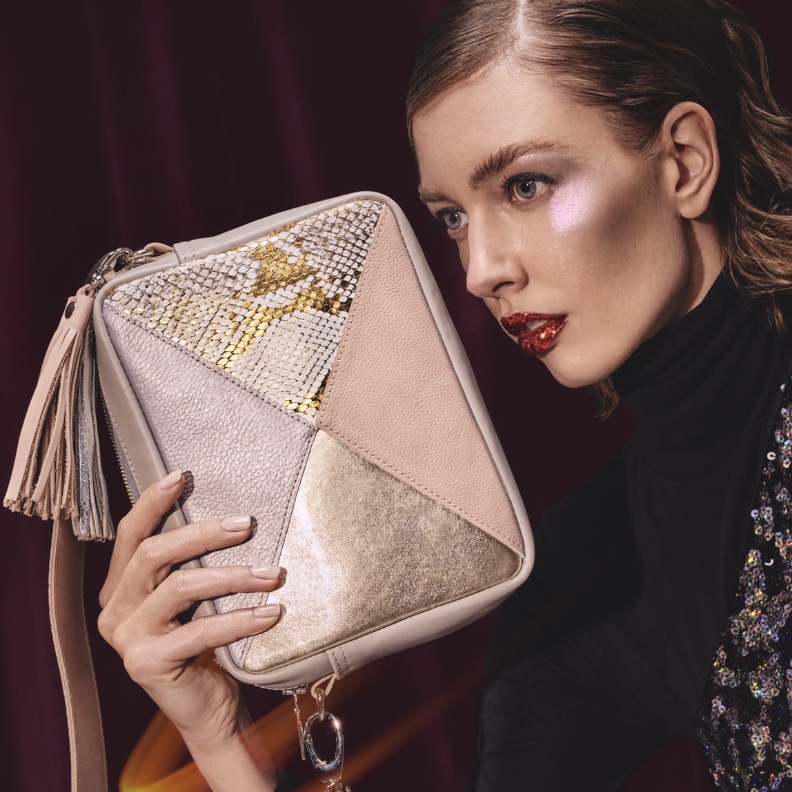 Model mit glänzendem Make-up präsentiert eine modische Clutch mit geometrischen Elementen aus verschiedenen Metallic-Tönen und Lederakzenten, perfekt für den eleganten Abendlook, präsentiert auf Cosmopolka.