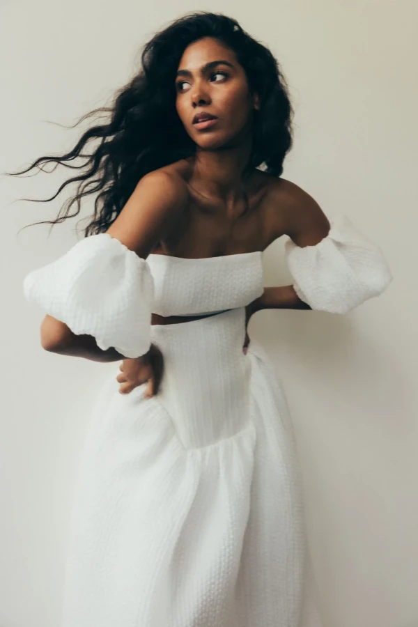 Moderne Braut in einem strukturierten weißen Brautkleid mit voluminösen Ärmeln, dynamische Pose und selbstbewusster Blick, vor minimalistischem Hintergrund.