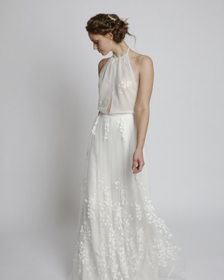 Eine Braut im langen Hochzeitskleid mit feinen Blumenstickereien steht vor einem weißen Hintergrund.