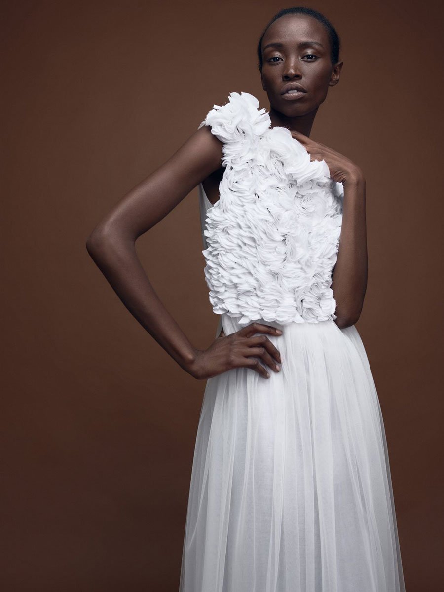 Ein weibliches Model in einem ärmellosen Brautkleid mit Rüschen steht vor einem braunen Studiohintergrund.