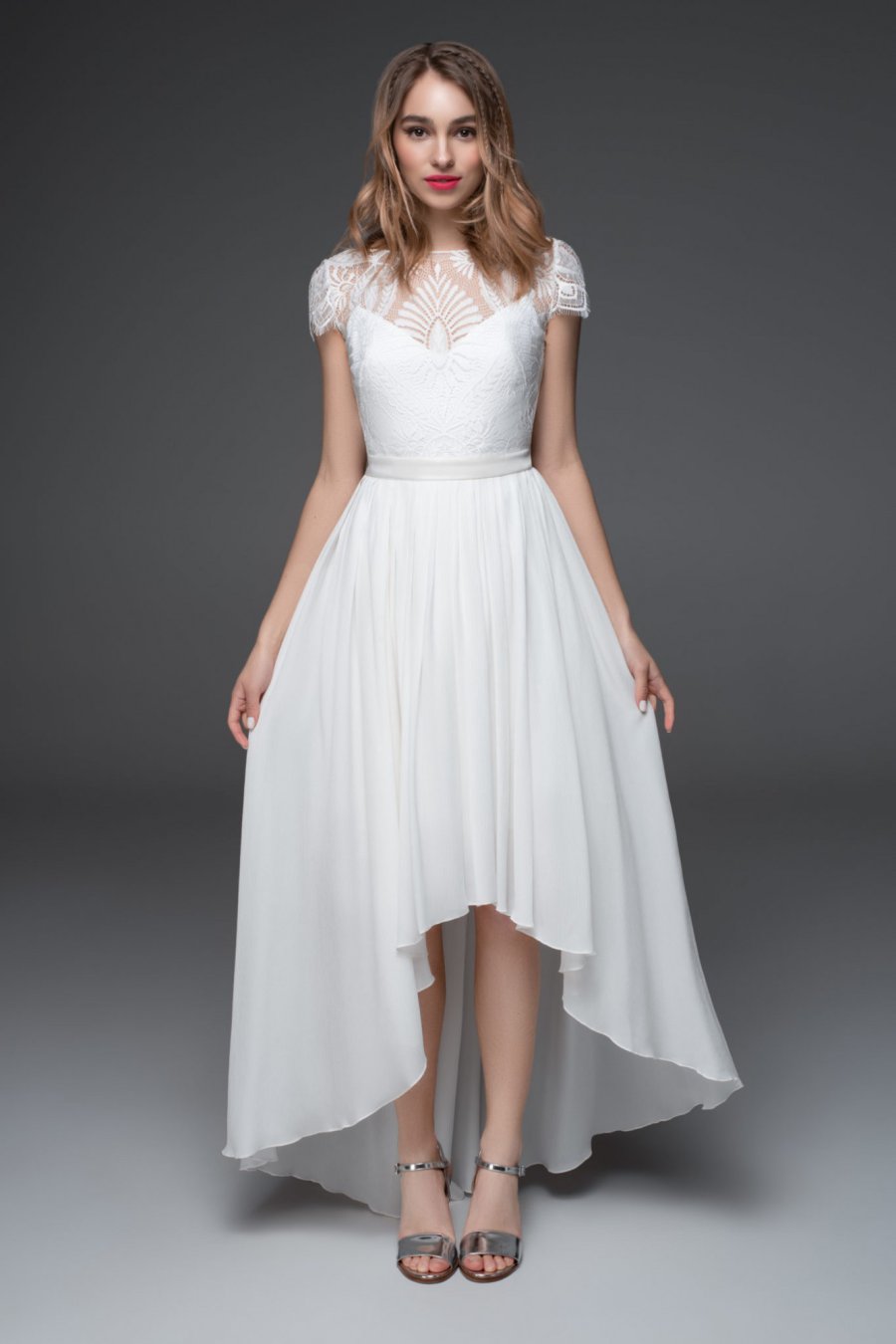Eine Braut trägt ein weißes langes Brautkleid mit und posiert vor grauem Hintergrund im Studio.