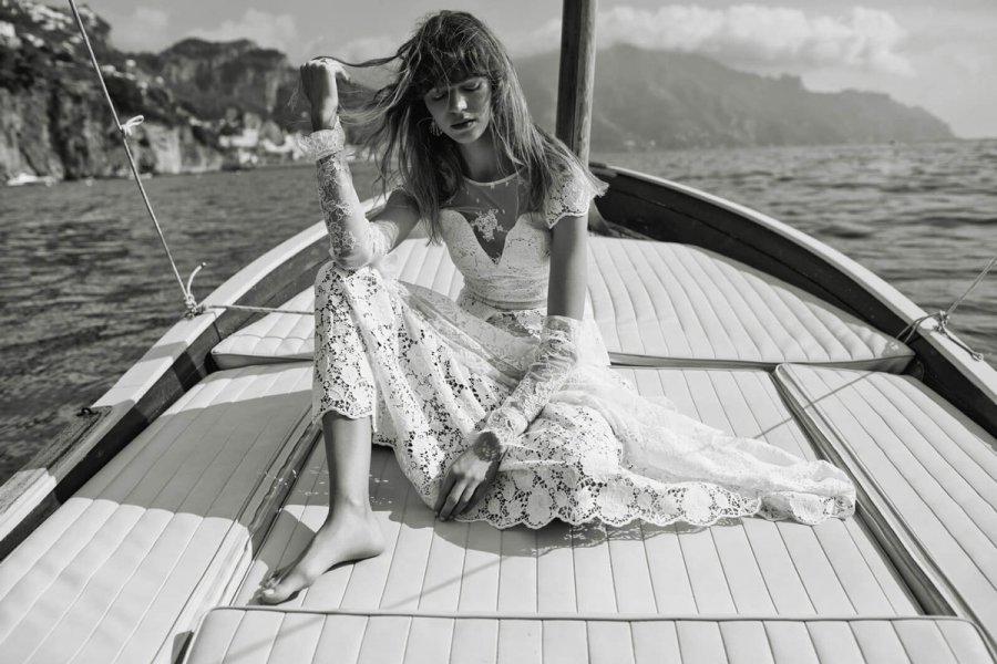 Eine barfüßige Frau im langen Brautkleid aus Spitze sitzt auf dem Deck eines Bootes, das sich vor einer felsigen Küste befindet.