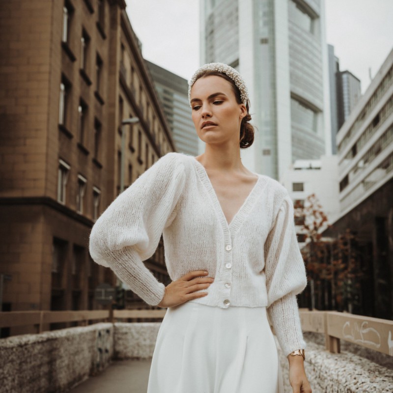 Braut posiert in einem weißen Braut-Cardigan mit V-Ausschnitt und langen Puffärmeln. Der Cardigan wird zu einem fließenden weißen Rock getragen, während im Hintergrund urbane Architektur sichtbar ist.