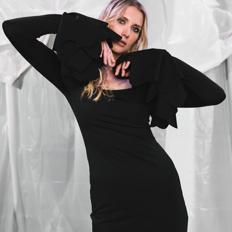 Model in schwarzem Midikleid mit weiten Ärmeln, posiert mit erhobenen Armen vor einem drapierten Hintergrund. Das Kleid betont eine elegante, figurbetonte Silhouette und künstlerische Ausstrahlung.