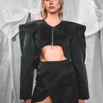 Schwarze Designer-Jacke mit breiten Schultern und asymmetrischem Rock – Model steht frontal mit intensivem Blick in die Kamera und betont die markante Silhouette der Jacke. Die kastige Passform und der minimalistische Schnitt verleihen dem Outfit einen modernen, avantgardistischen Look. Fotografiert vor silbernem Stoffhintergrund. thumbnail