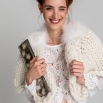 Eine Frau in einem cremefarbenen, dicken Braut-Cape aus Bio-Wolle, ergänzt durch einen Marabou-Federkragen und ein weißes Spitzen-Top. Ein lässig-eleganter Look für die stilbewusste Braut. thumbnail