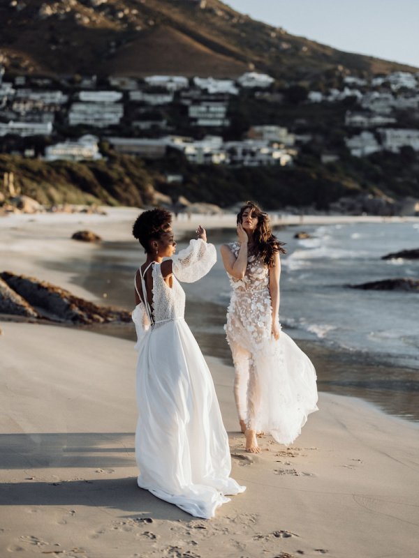 Zwei Bräute in Boho-Brautkleidern von Kisui Berlin spazieren barfuß am Strand entlang, die fließenden Kleider mit Spitze und Blumenmustern fangen die unbeschwerte Stimmung des Augenblicks ein.