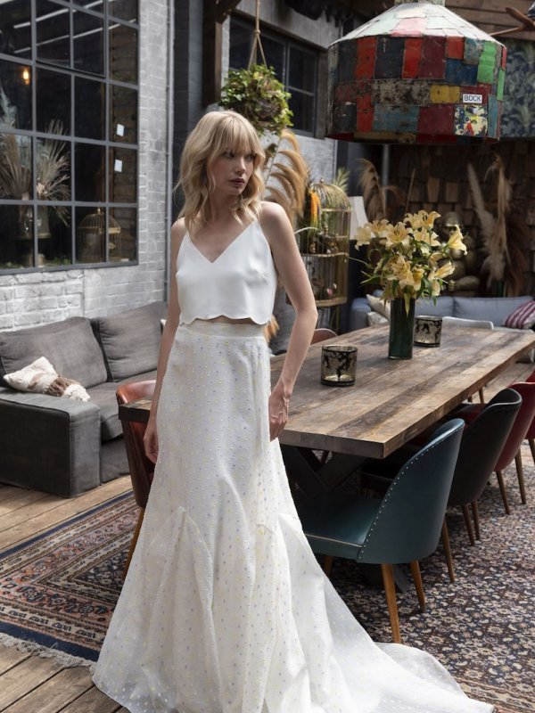 Vintage Brautkleid von Kisui Berlin, in einem eleganten, rustikalen Raum mit hölzernem Tisch und Pflanzen im Hintergrund, getragen von einer Braut, die entspannt in einem lichtdurchfluteten, klassischen Ambiente steht.