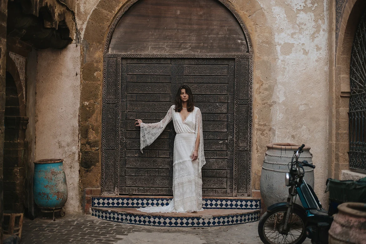 Elegantes Boho-Brautkleid Iga aus der Kollektion von Karolina Twardowska, getragen von einer Braut vor einer verzierten, historischen Holztür in einer rustikalen Altstadt-Kulisse. Das Kleid besticht durch seinen fließenden, spitzenbesetzten Stoff und weite Ärmel im Vintage-Stil.