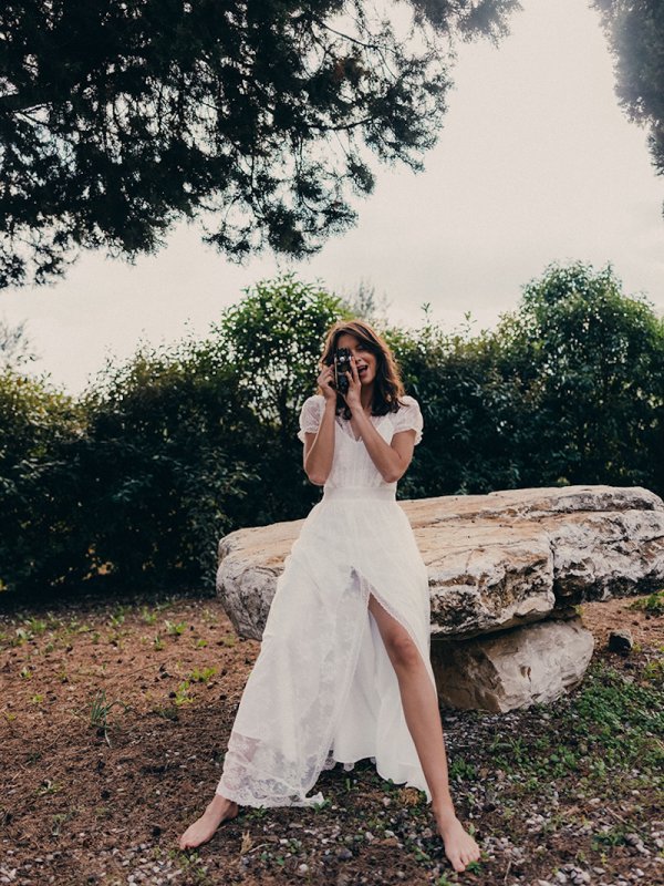 Boho Brautkleid von Karolina Twardowska, mit zarter Spitze und einem hohen Beinschlitz, getragen von einer Braut, die barfuß auf einem großen Stein sitzt und fotografiert, umgeben von natürlicher Vegetation und Bäumen.