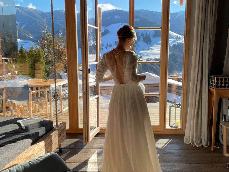 Braut in einem rückenfreien Kleid in einem Chalet mit Bergblick – Eine Braut steht in einem Chalet mit einer atemberaubenden Aussicht auf die verschneiten Berge, sie trägt ein fließendes, rückenfreies Hochzeitskleid, ideal für eine winterliche Berghochzeit.