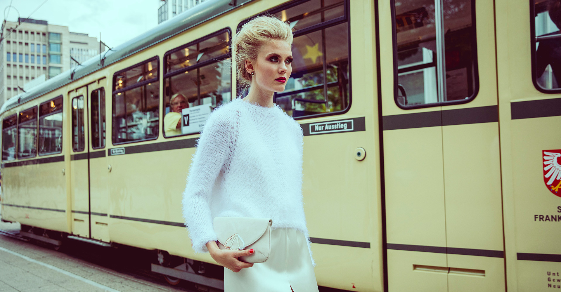 Moderne Braut in einem eleganten weißen Pullover und Rock, posierend vor einer historischen Straßenbahn in urbaner Kulisse. Brautmode-Design von Marryandbride.